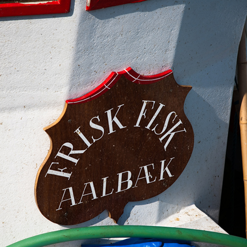 Skilt af træ med teksten ‘Frisk fisk Aalbæk’, monteret på en lys væg